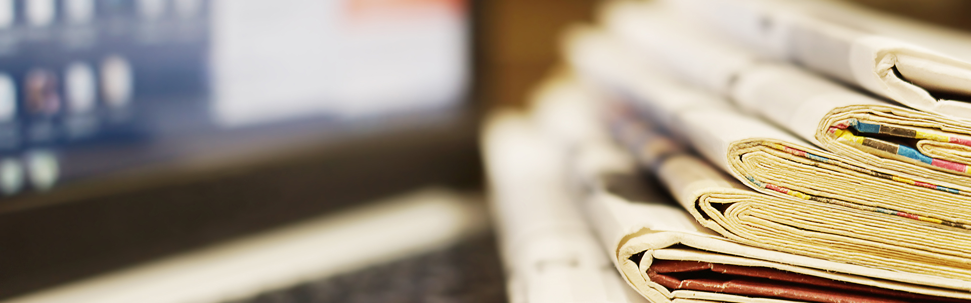 Close-up of neatly stacked newspapers and magazines, showcasing the variety of content available, with a blurred computer screen in the background.